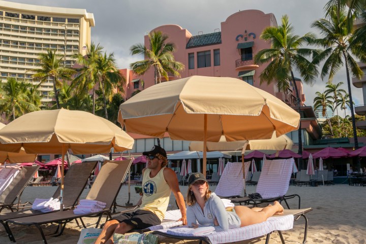 Two people relax on beach loungers under umbrellas near a pink building and palm trees.