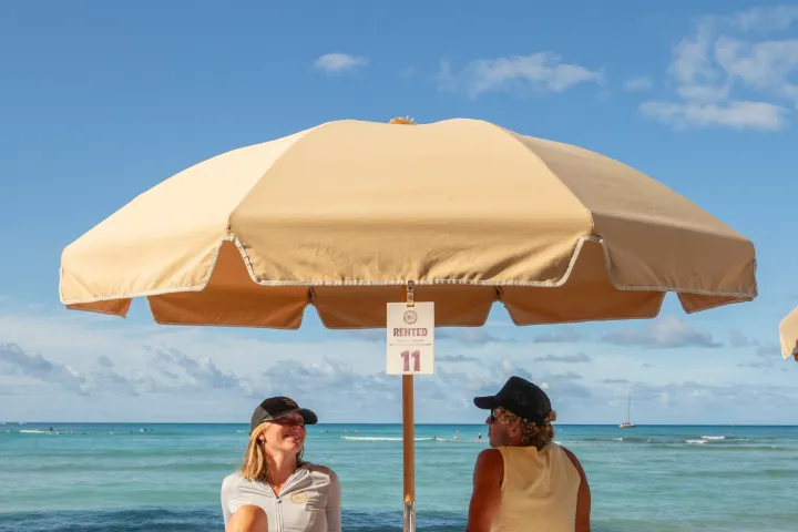 Two people relax on beach chairs under an umbrella, with a blue ocean in the background.