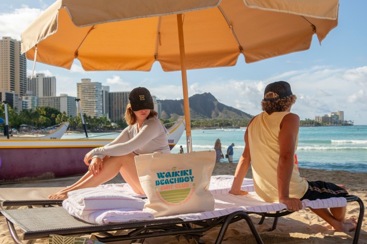 Two people sit under an umbrella on a beach, with city skyline and ocean in the background.