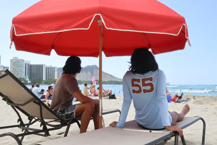 Two people sit on lounge chairs under a red umbrella on a sandy beach with city buildings in the background.