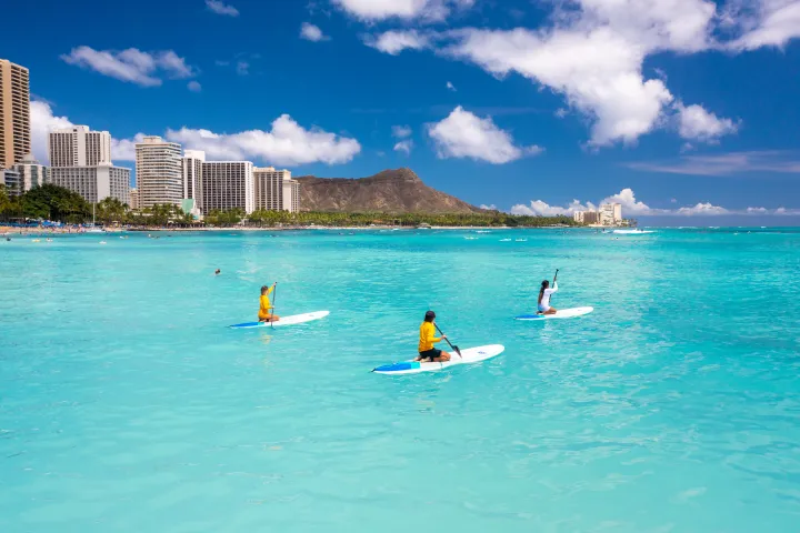 Three people paddleboarding on turquoise ocean with city and mountain in background.