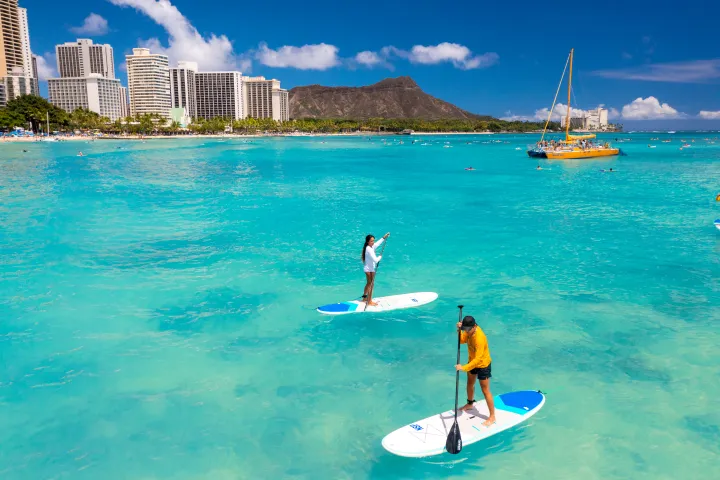 Two people paddleboarding on a turquoise ocean with city buildings and a mountain in the background.