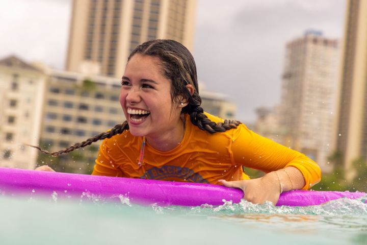 Woman in orange rash guard smiling on a pink surfboard in the water, city buildings in the background.