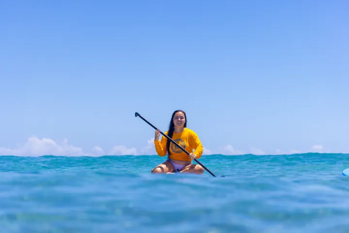 Person in a yellow shirt stand-up paddleboarding on clear blue water under a bright sky.