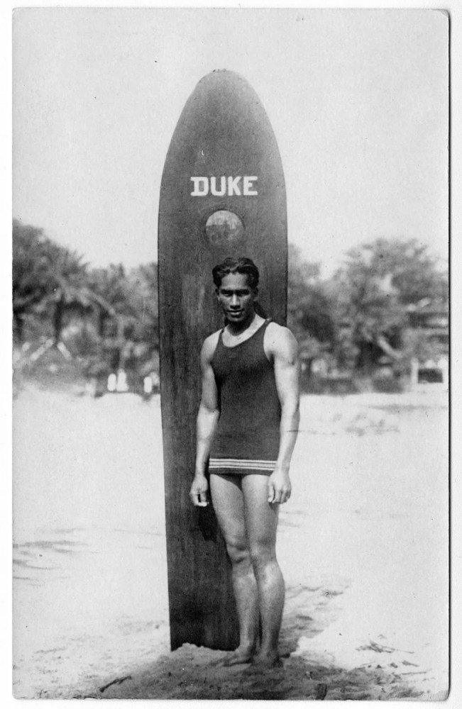 Person in a swimsuit stands beside a surfboard labeled 'DUKE' on a beach.