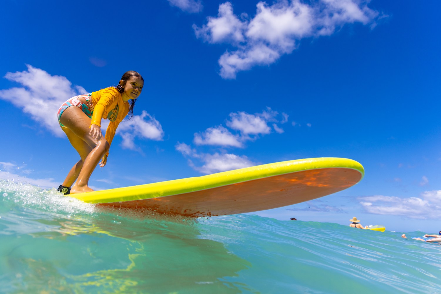 Smiling person surfing on a yellow board in clear blue ocean under a sunny sky.