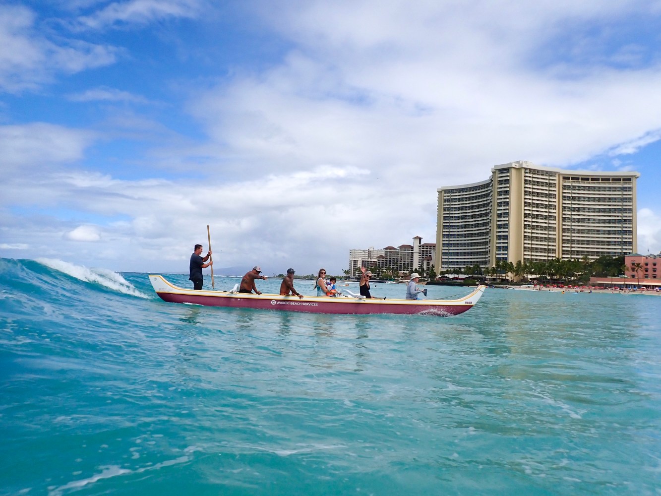 Group paddling an outrigger canoe in ocean near a large hotel under a cloudy sky.
