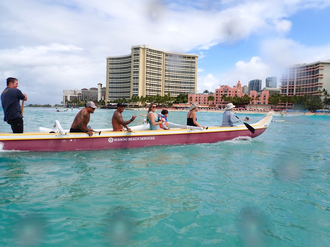 People in a canoe paddling on the ocean with tall buildings in the background.