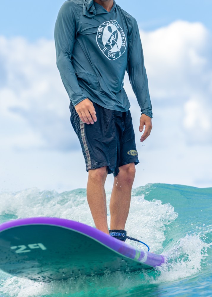 Person surfing on a purple board, wearing a gray shirt and black shorts, on a wave.