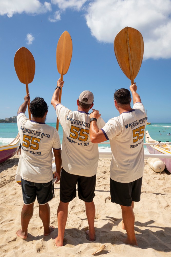 Three people on a beach holding paddles, facing the ocean.