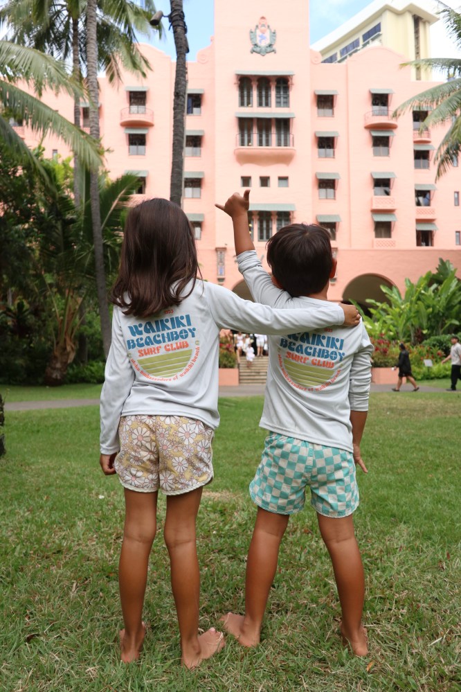 Two children in matching shirts face a pink hotel, one raising an arm, surrounded by palm trees.
