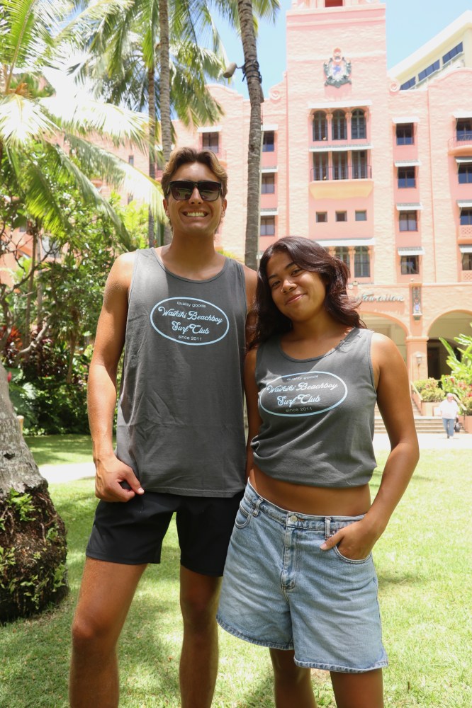 Two people in matching gray tank tops smile in front of a pink building with palm trees.