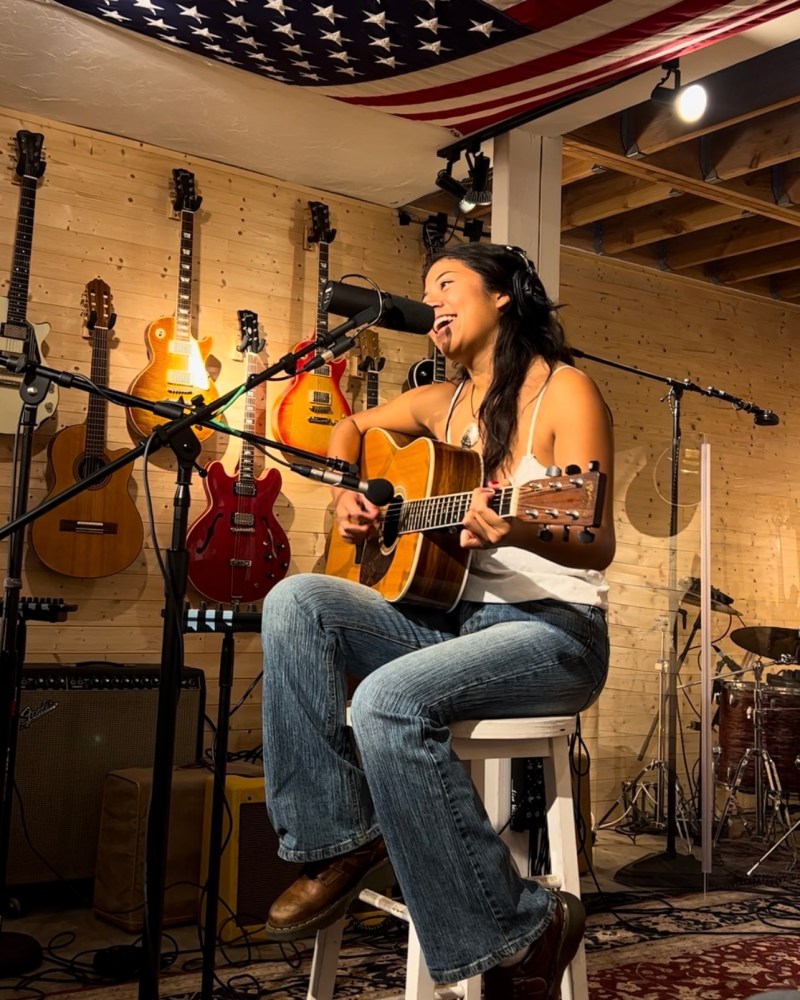 Person playing acoustic guitar and singing in a studio with guitars and drums.
