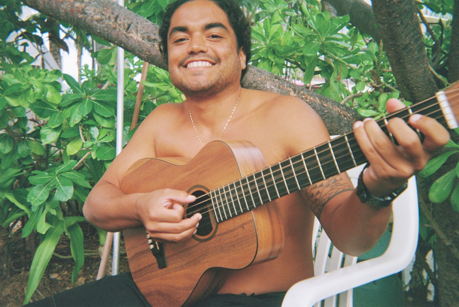 Smiling person sitting outdoors, playing an acoustic guitar.