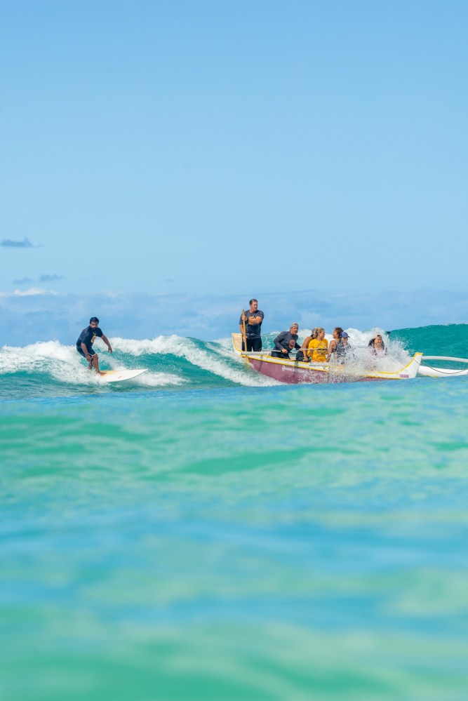 Surfer rides a wave beside a canoe with people, under a clear blue sky.