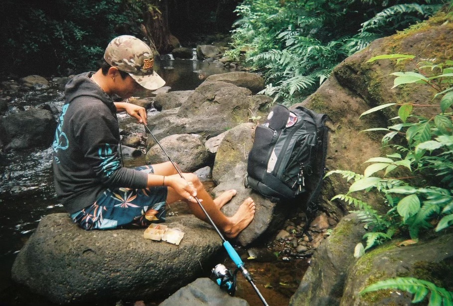 Person sitting on rocks by a stream, holding a fishing rod, backpack nearby.