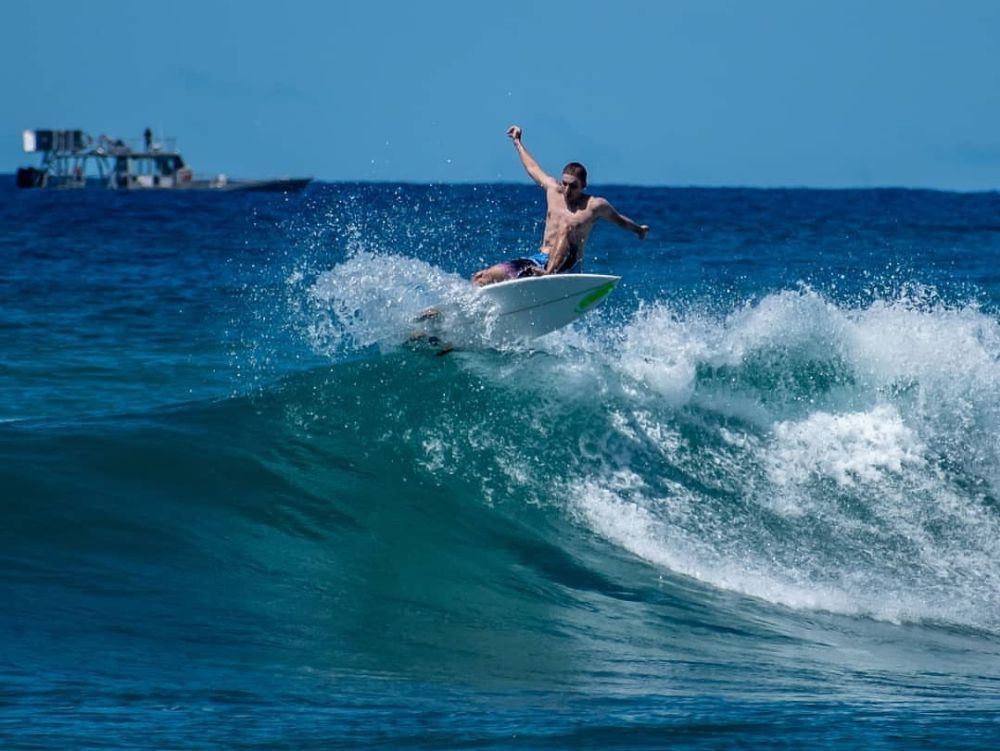 Surfer riding a wave with a ship in the background under a clear blue sky.