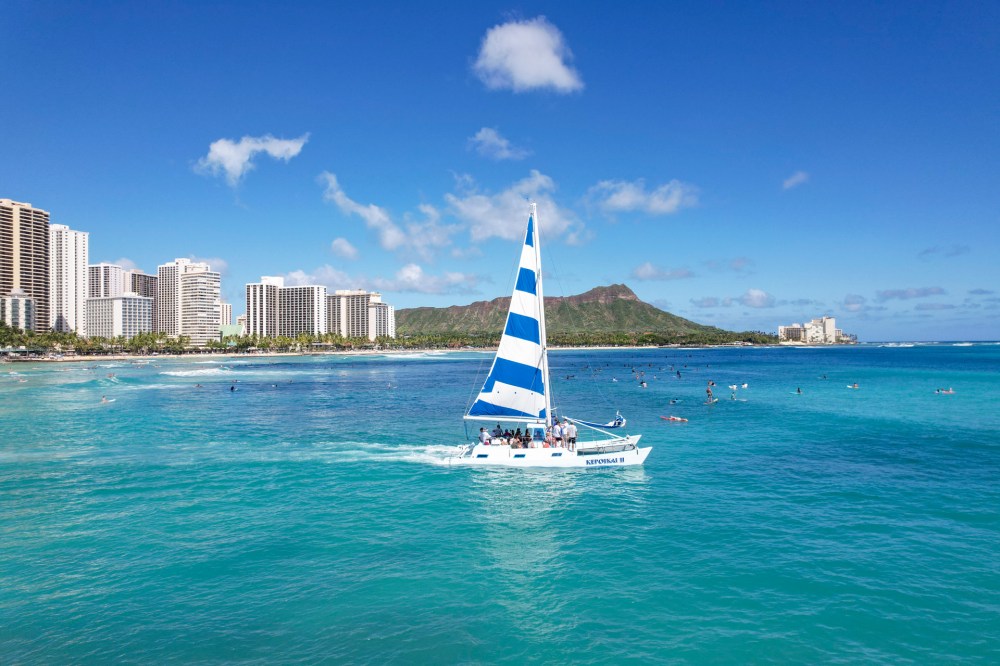 Sailboat with blue and white stripes on turquoise water near city skyline and mountain.