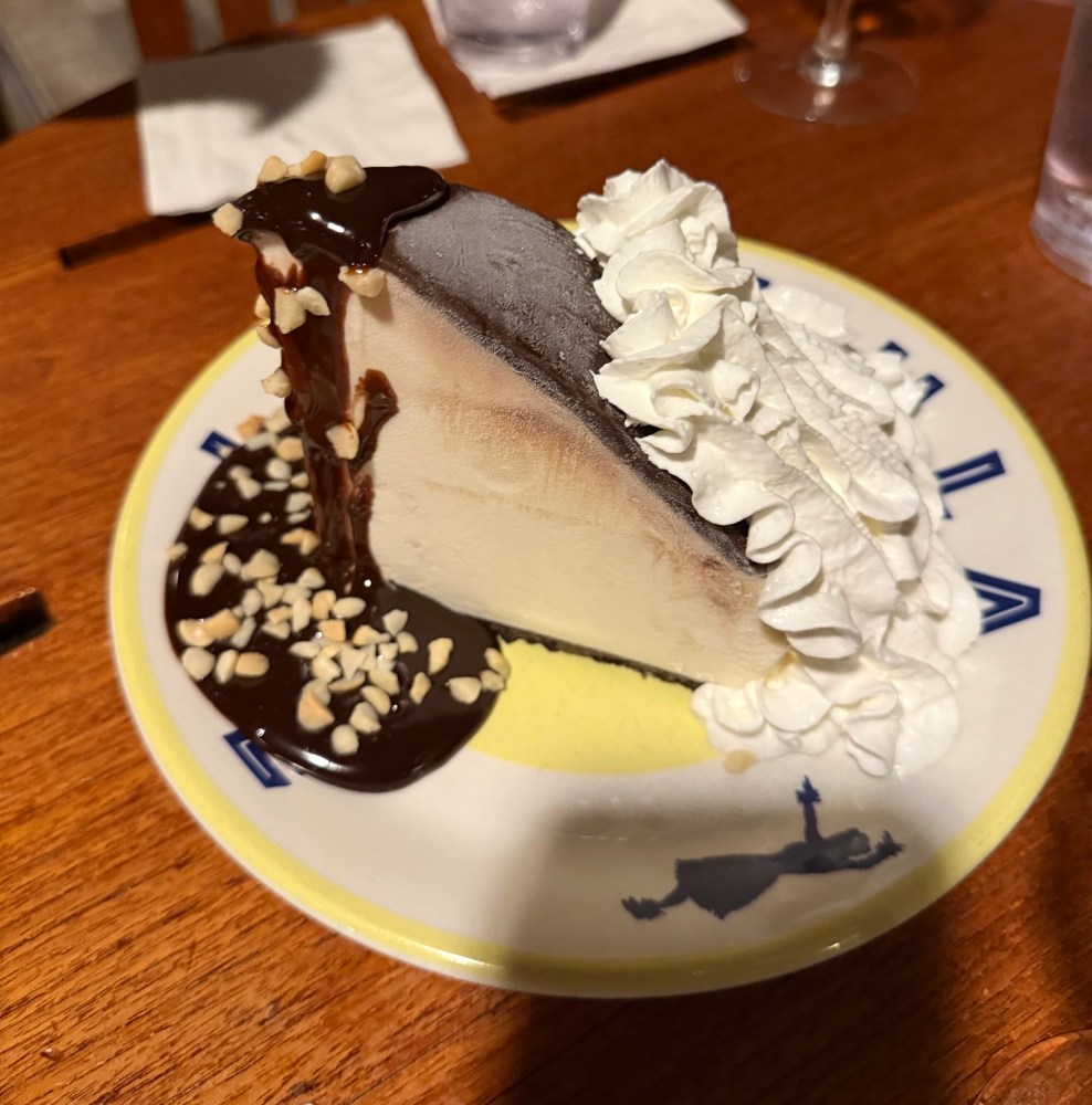 Slice of cheesecake with chocolate sauce, nuts, and whipped cream on a decorative plate.