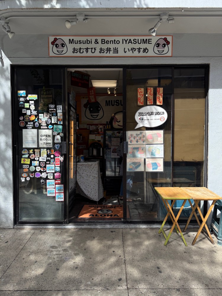 Small restaurant entrance with signs and stickers, featuring a wooden table outside.
