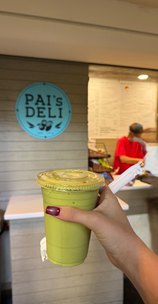 Hand holding a green smoothie in front of a deli counter.