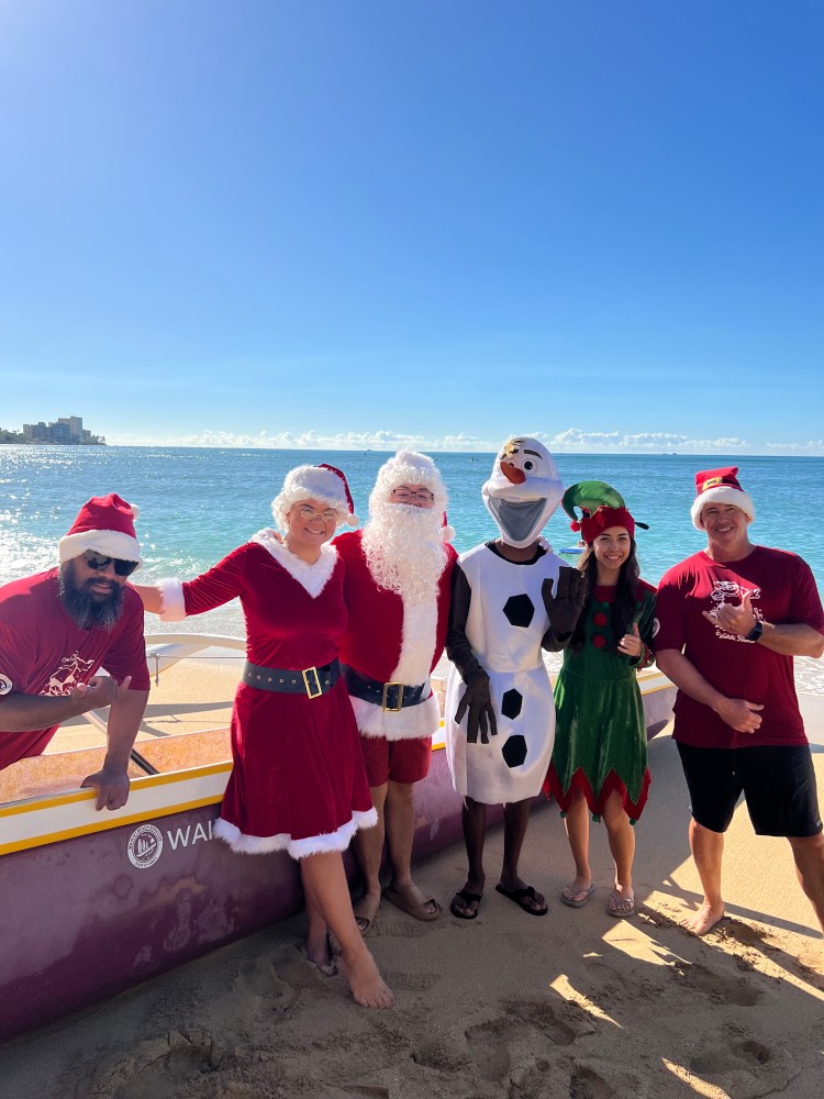 People in festive outfits with Santa and snowman costumes on a beach near a canoe.