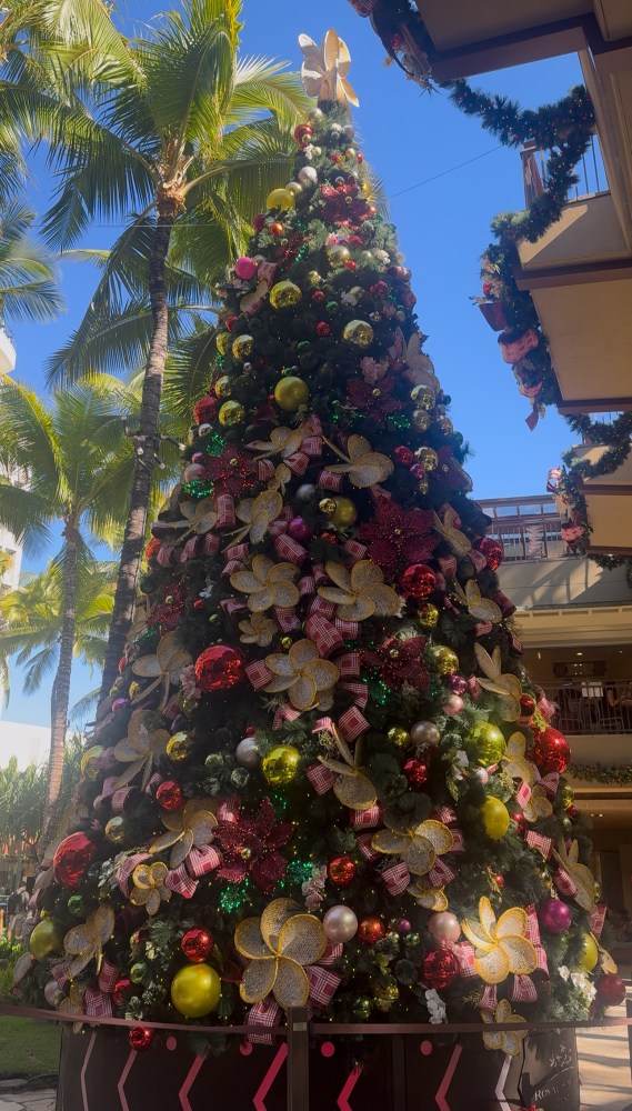 Large decorated Christmas tree with colorful ornaments under palm trees in a mall.