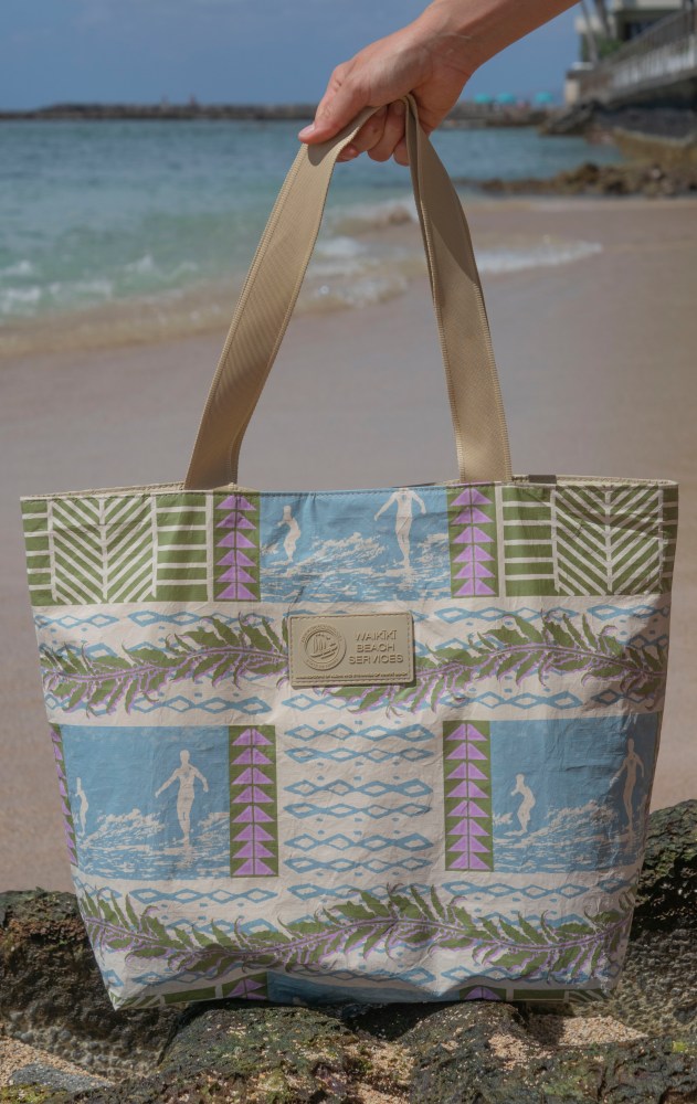 Hand holding patterned beach tote with 'Waikiki Beach Services' label on sandy shore.