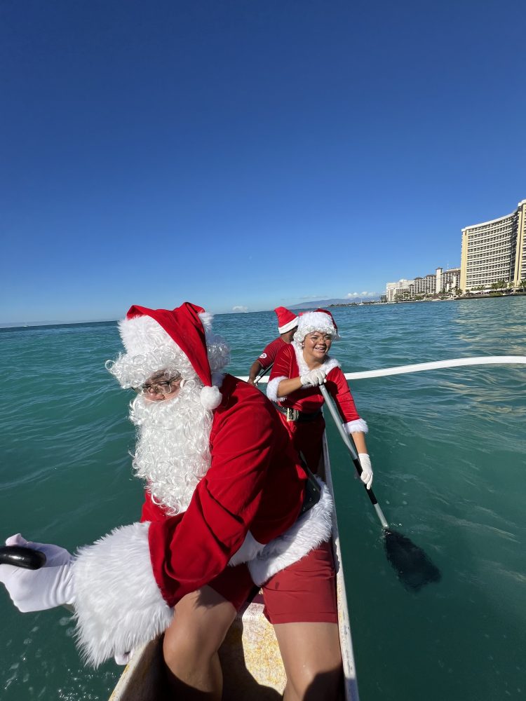 People dressed as Santa paddling a boat in blue ocean with city buildings in background.