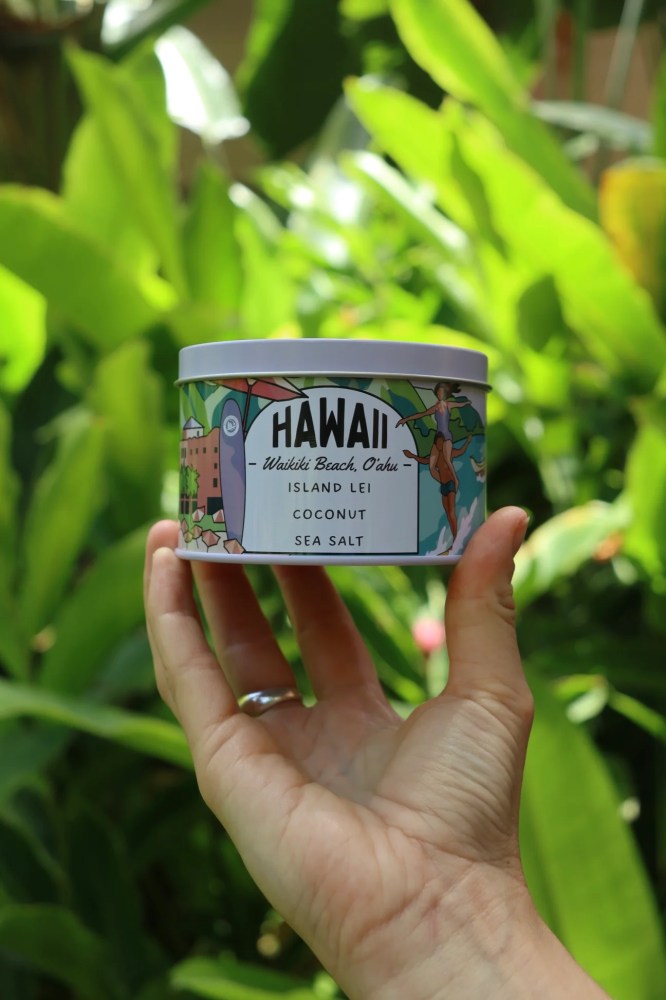 Hand holding a Hawaii-themed candle tin with lush green background.