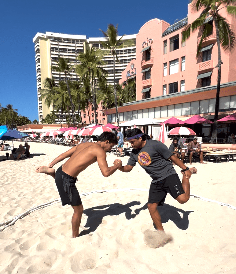 Two men on a beach playing a game, holding one foot and clasping hands.