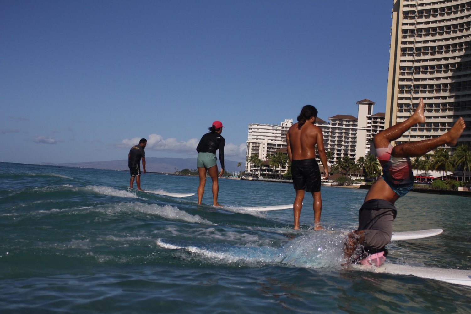 Four people surfing near a city shore, one person falling off their board into the water.