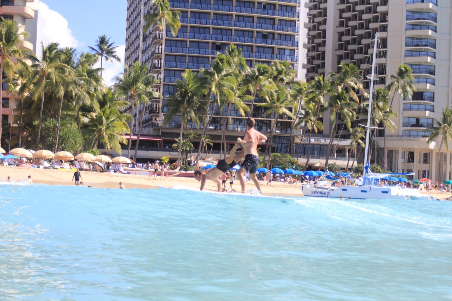Beach scene with people playing in water, buildings, and palm trees in background.