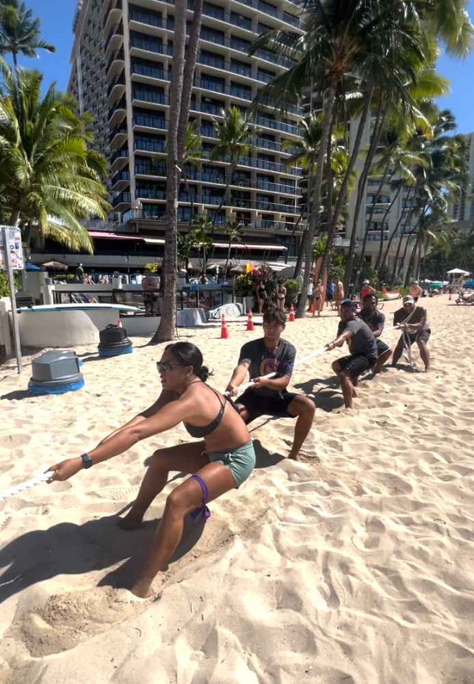 People playing tug-of-war on a sandy beach with buildings and palm trees in the background.