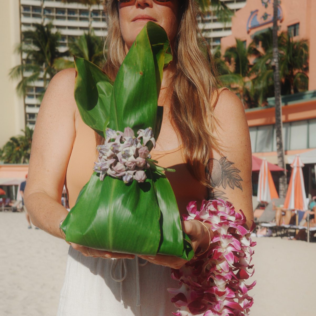 Woman on beach holds a green leaf bundle and purple flower lei.