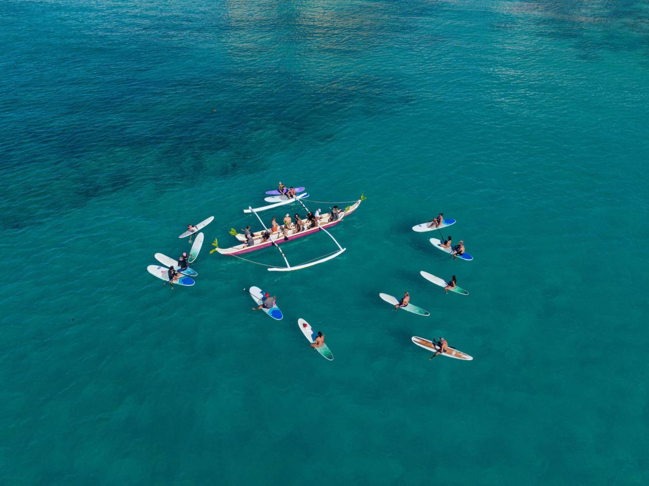 A group of people on surfboards and a canoe in blue ocean water, viewed from above.