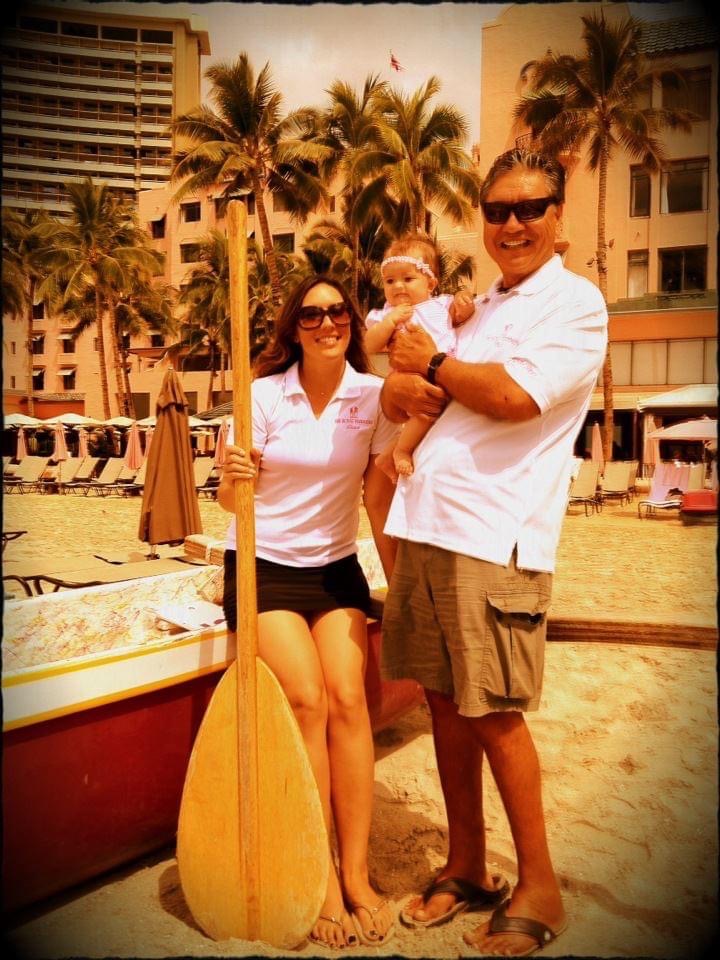 Family with a baby posing on a beach with palm trees and hotel in the background.