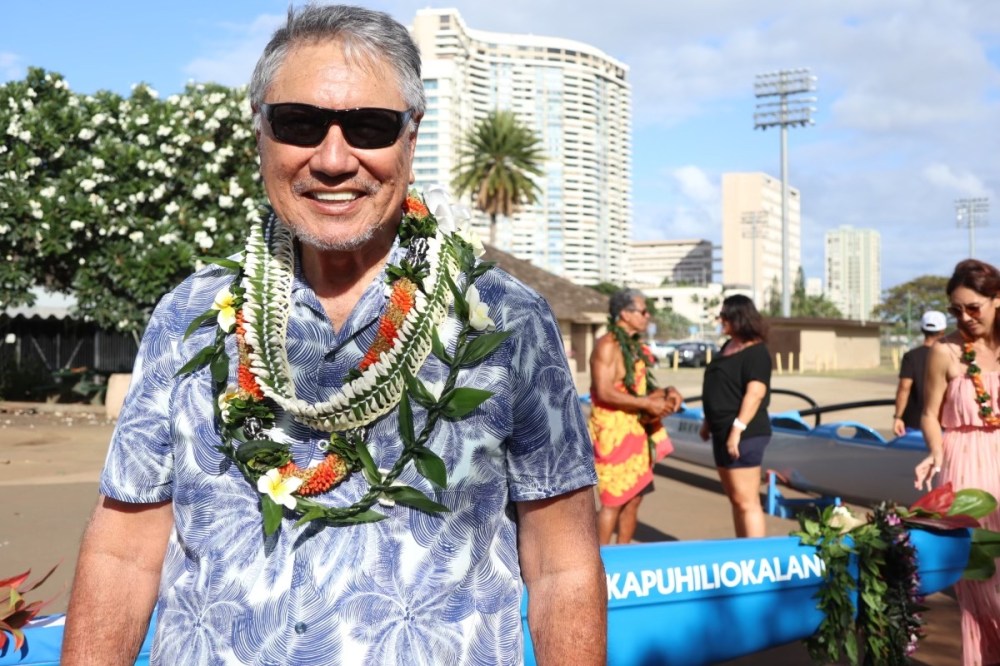 Smiling man in sunglasses with flower lei, palm trees, and people in background.