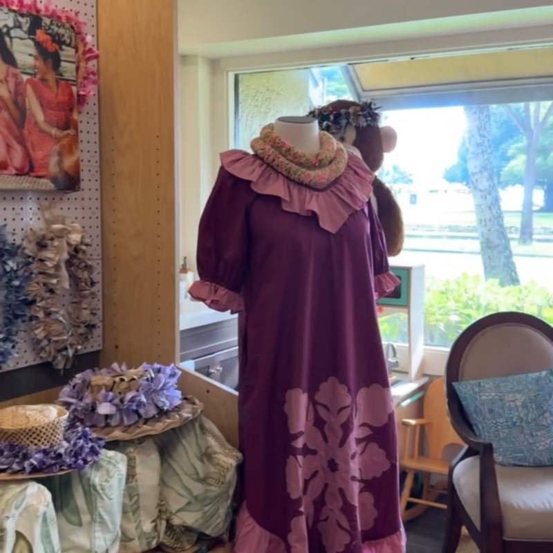 Mannequin in purple dress with floral patterns and leis in a storeroom with textiles and decor.
