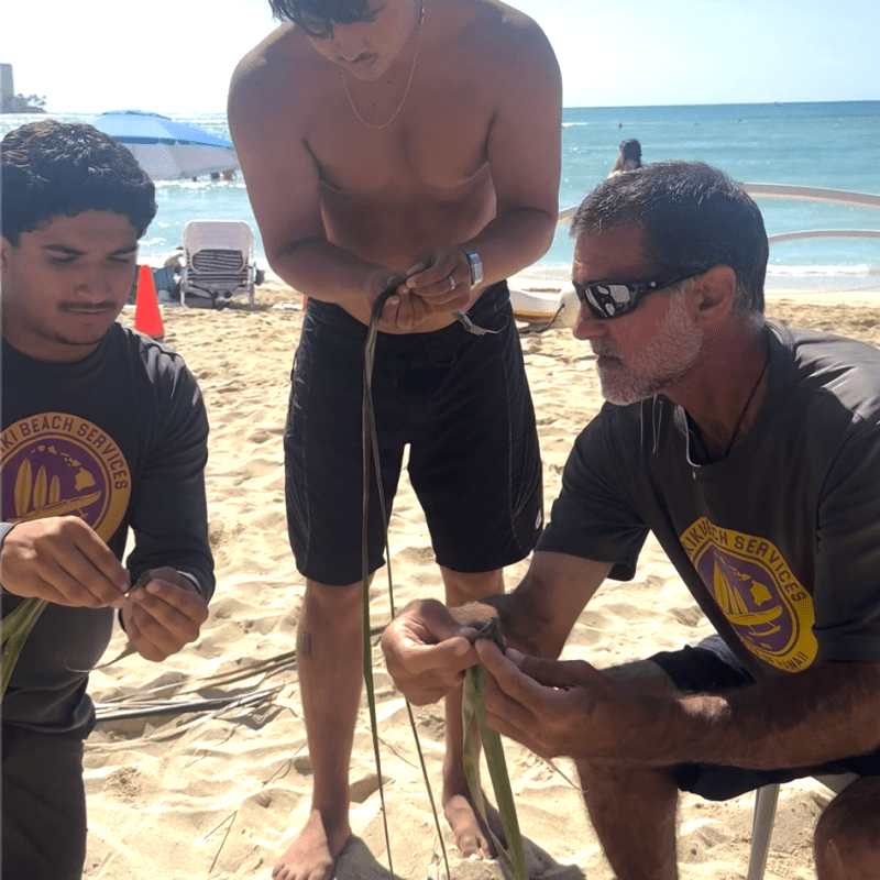 Three men on a sunny beach weaving long leaves, two sitting and one standing.