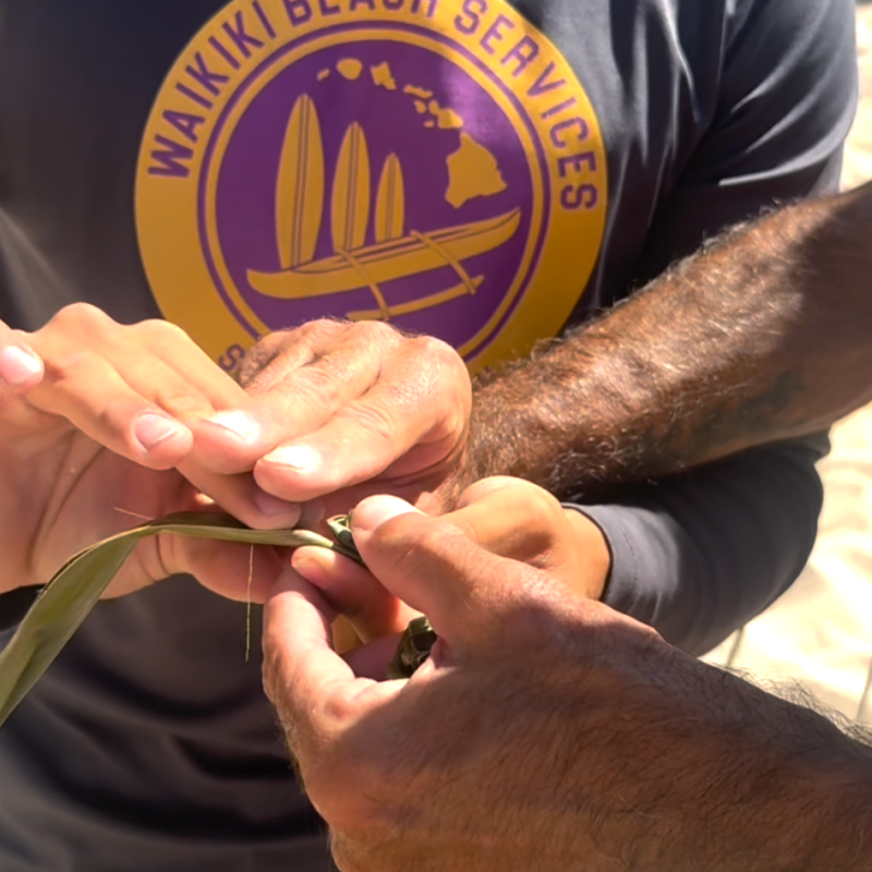 Close-up of hands weaving a palm leaf, with a 'Waikiki Beach Services' shirt in the background.