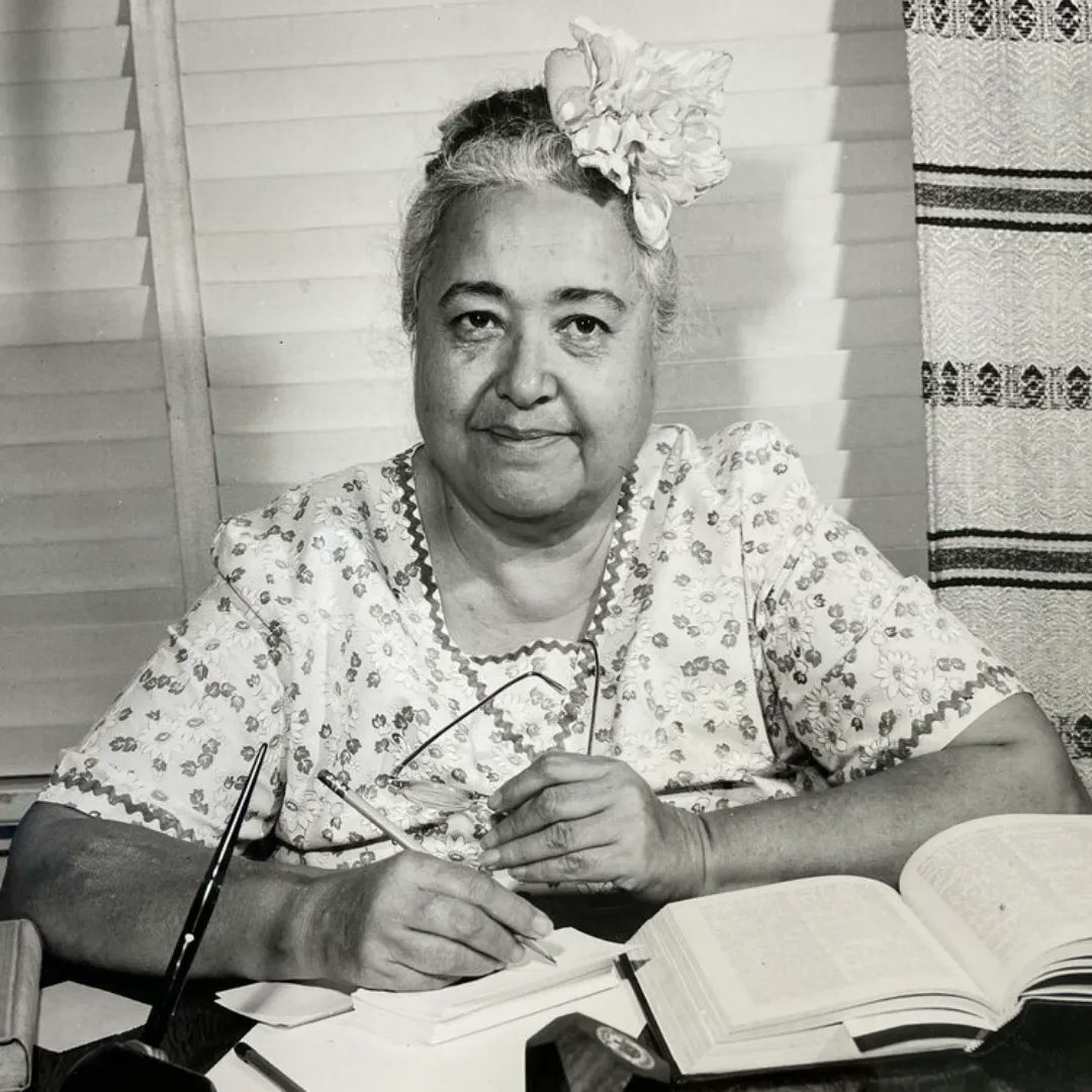 Woman with flowers in her hair writing at a desk with open books.