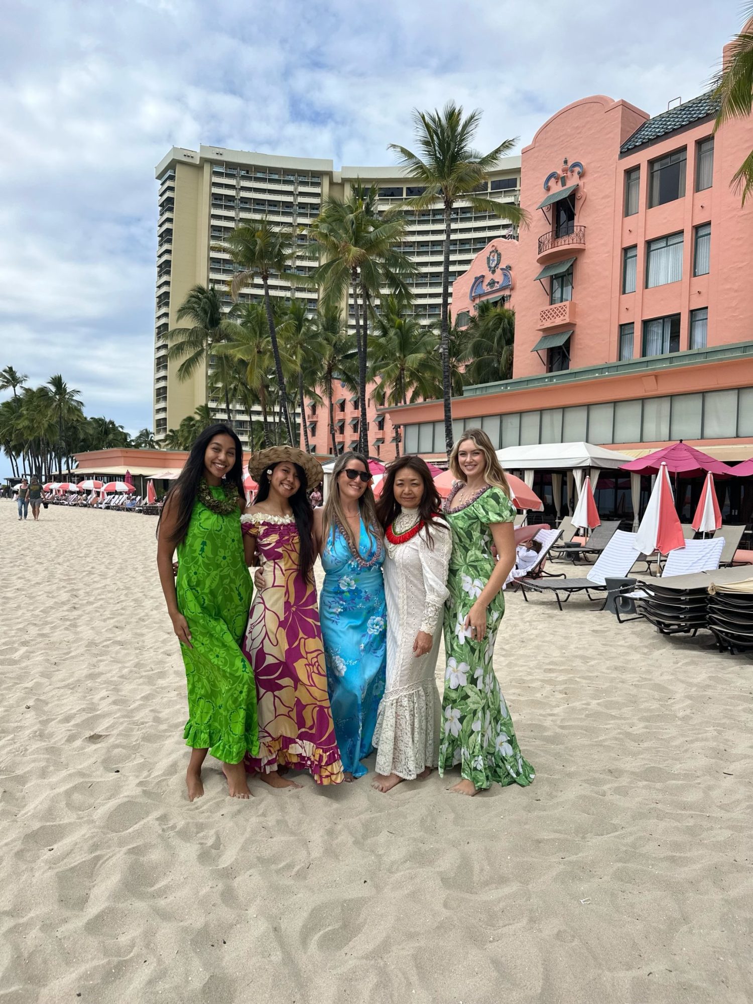 Five women in colorful dresses stand on a beach with a pink hotel and palm trees in the background.