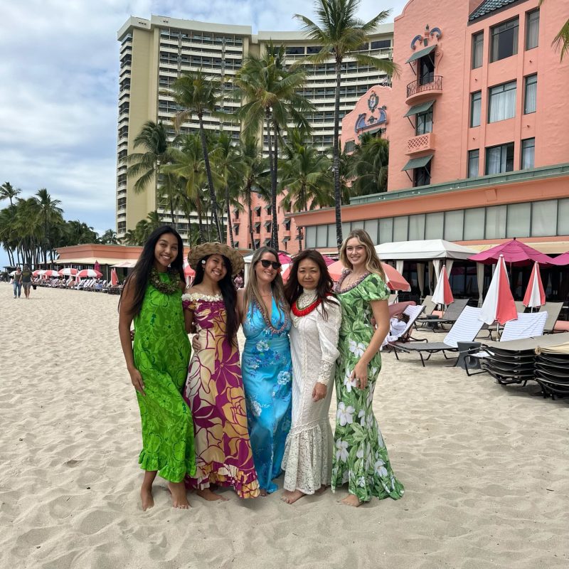 Five women in colorful dresses stand on a beach with a pink hotel and palm trees in the background.