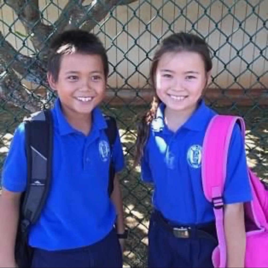 Two smiling children in blue uniforms with backpacks outside near a fence and tree.
