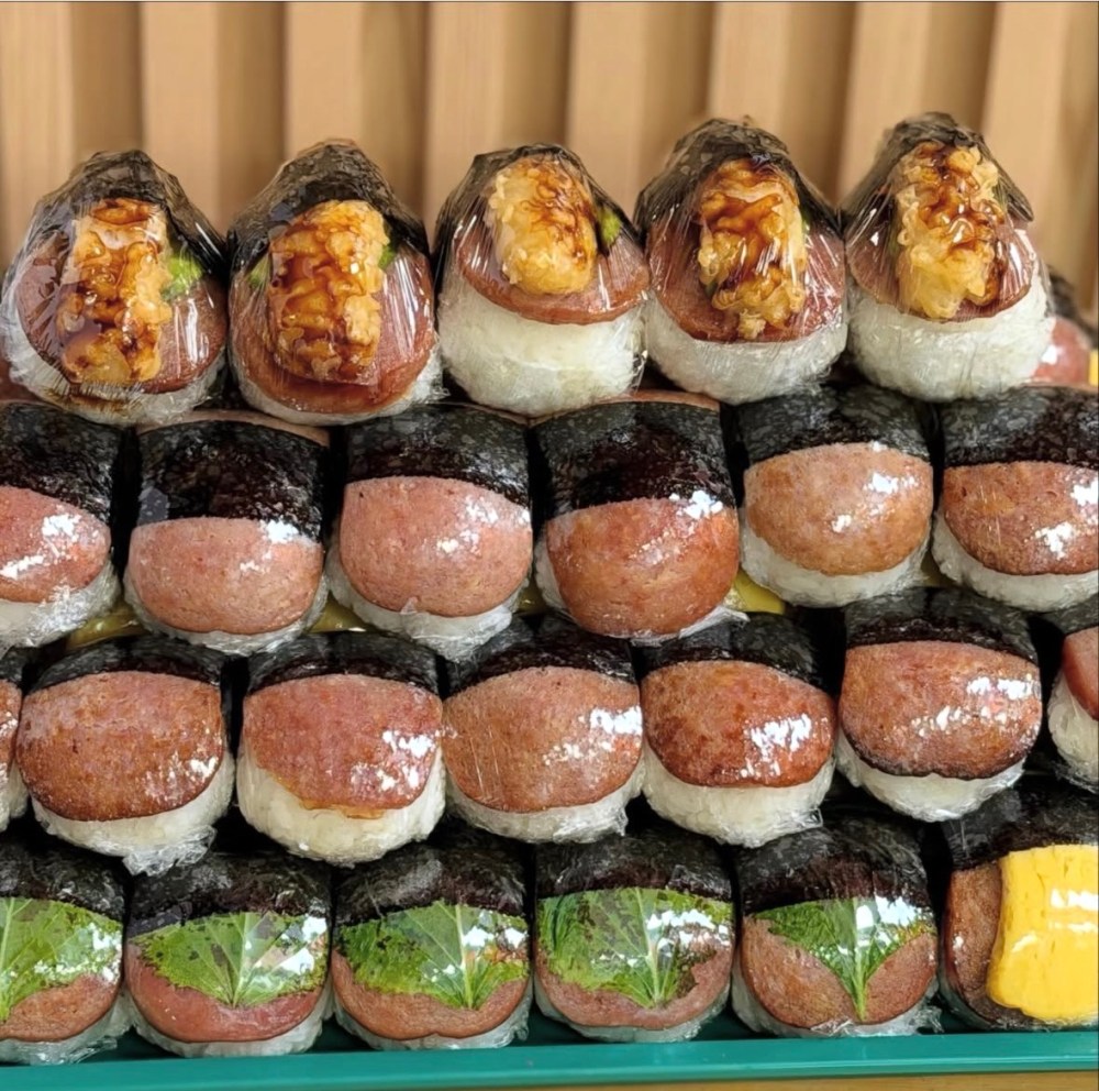 Stack of various musubi wrapped in plastic with different toppings visible.