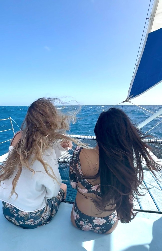 Two women with flowing hair sit on a boat under a blue sky and sail.