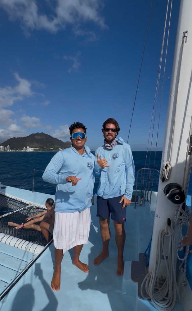 Two people on a boat in blue shirts, showing shaka signs with ocean and mountain in background.