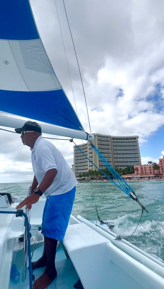 Person steering sailboat with blue sail near tall building and cloudy sky.