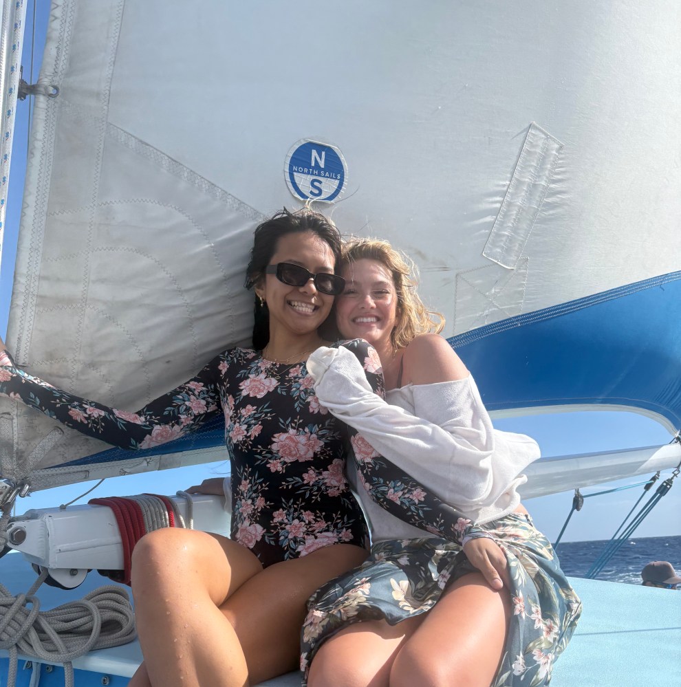 Two women smiling on a sailboat, sitting by a sail, with a clear sky.